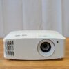 Compact white Optoma 4K projector rental unit with ventilation grilles and large zoom lens on a wood table