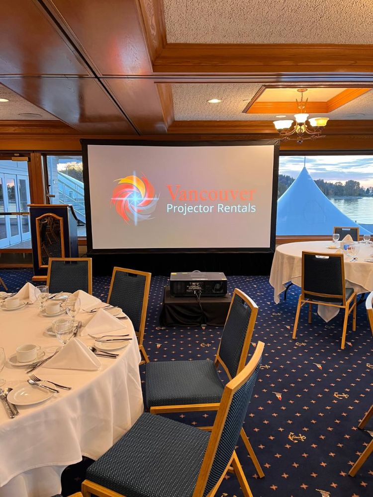 Large projection screen and projector rental in a formal dining event room with set tables and a scenic waterfront backdrop