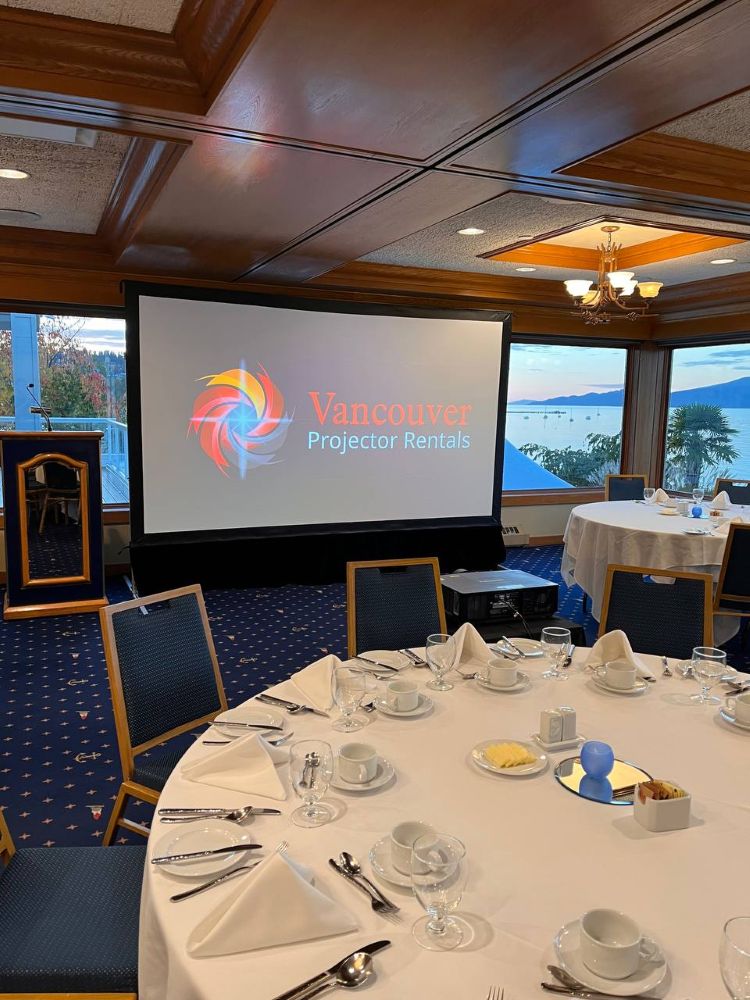 Wide-angle view of a banquet room with a large projection screen and projector rental, set up for a formal event with waterfront views