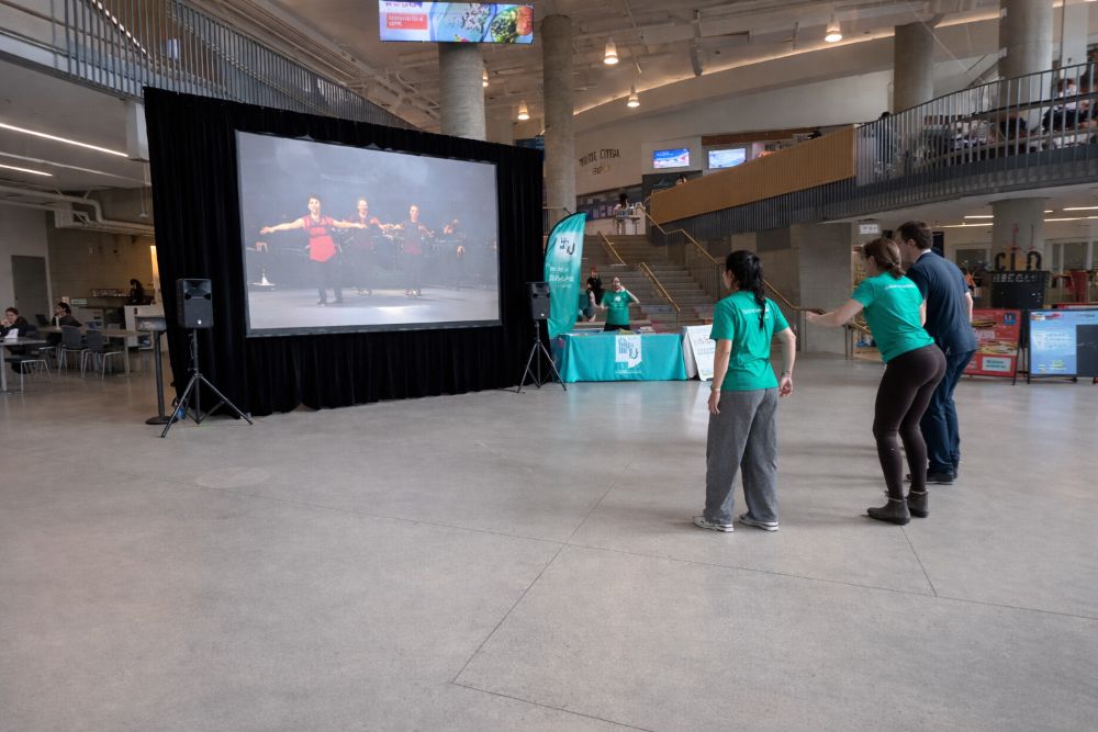 Large rear screen rental with black drape surround set up in a UBC campus atrium, with participants dancing in front of the projected video content