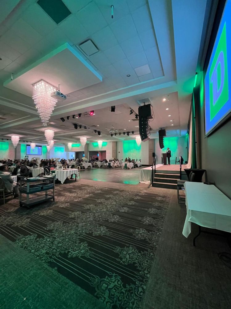 Side view of a TD Bank corporate gala with ceiling-mounted line array speakers, stage lighting rig, mirror balls, and a presenter at the podium