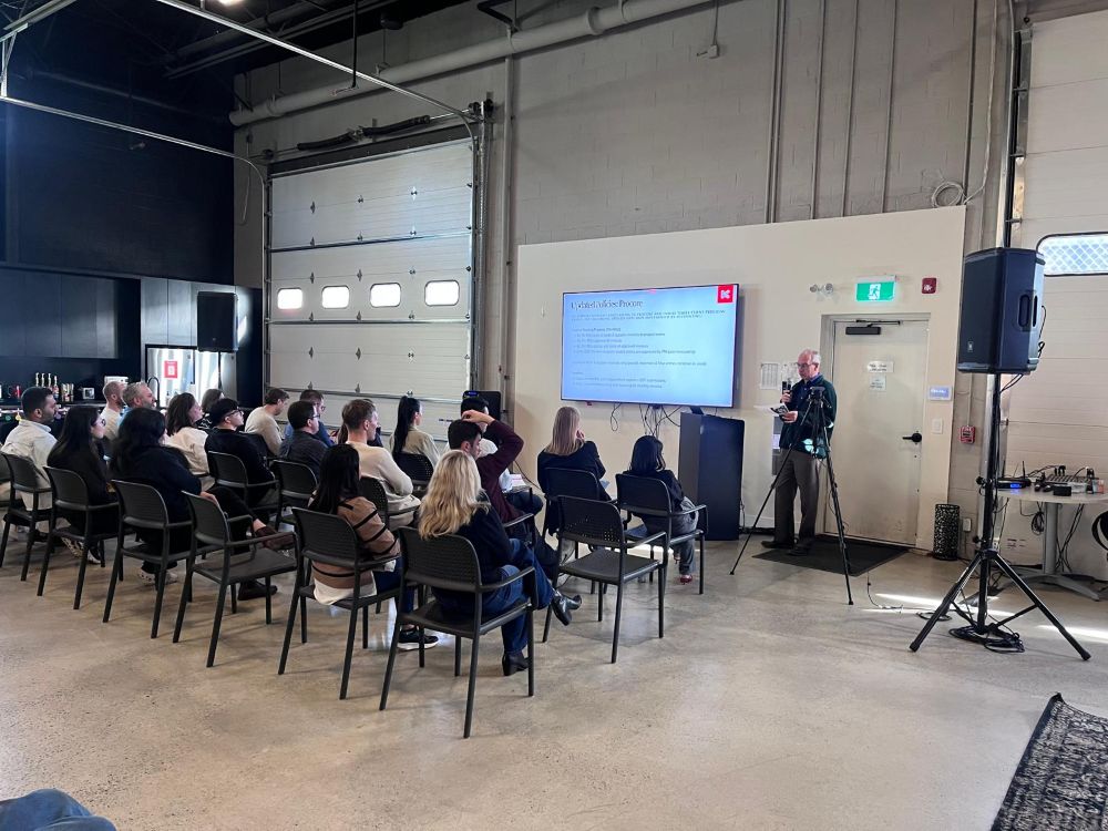 TV rental on a stand displaying a corporate presentation slide, with a speaker at a microphone and PA speaker on a tripod in an industrial warehouse venue
