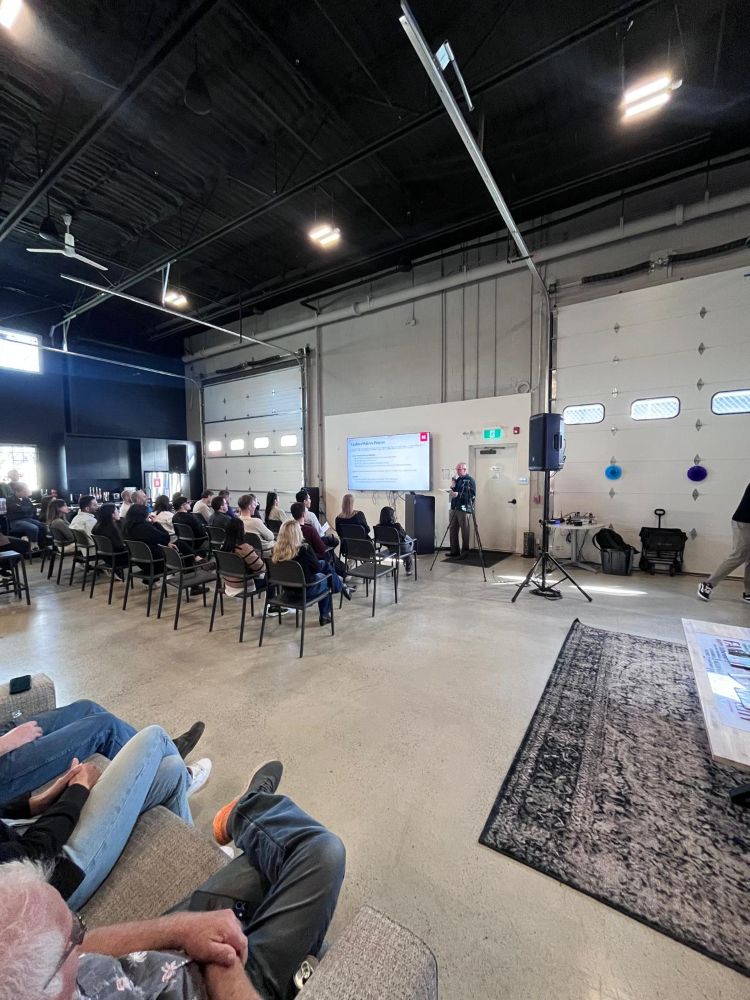 Wide-angle view of a corporate event in a large industrial warehouse with a TV rental, tripod-mounted PA speaker, and full seated audience