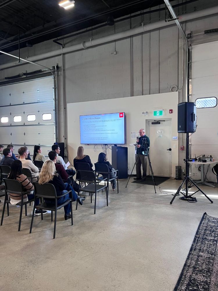 Full audience view of a corporate presentation in a warehouse venue with a TV rental on a stand, PA speaker on a tripod, and AV mixing table