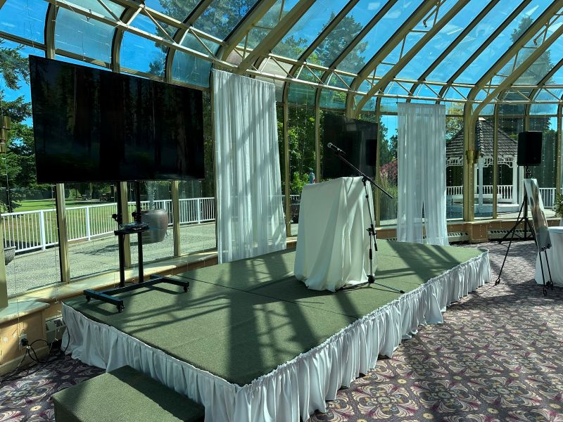 TV rental and microphone stand set up on a draped stage with white curtain backdrop in a sunlit glass conservatory event space